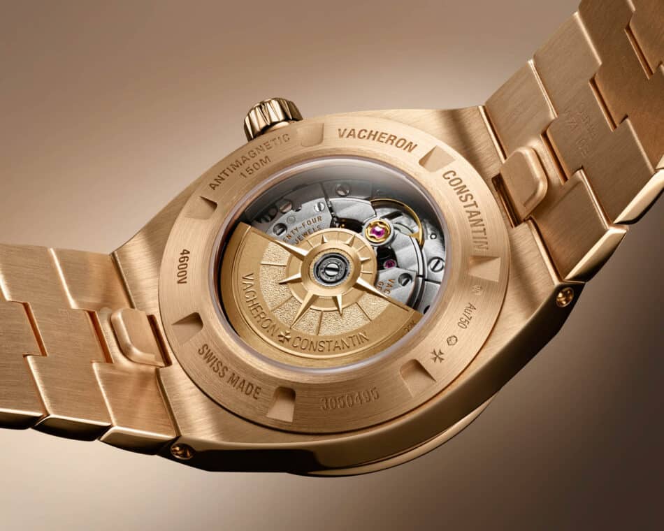 VACOverseasSelfWindingBACK4600V200RH128 Watches World Primer plano del fondo de un reloj de oro en el que se aprecia el mecanismo mecánico y el nombre de la marca Vacheron Constantin grabado con el brazalete metálico visible al fondo
