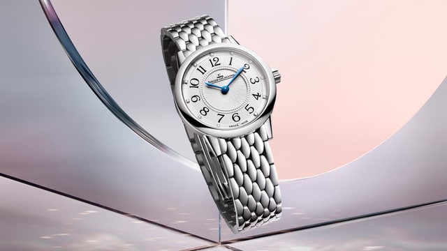 RendezVous HourMinute Watches World Reloj de pulsera plateado con brazalete de eslabones metálicos y esfera blanca con agujas azules que destaca sobre un fondo abstracto de tonos rosa y azul
