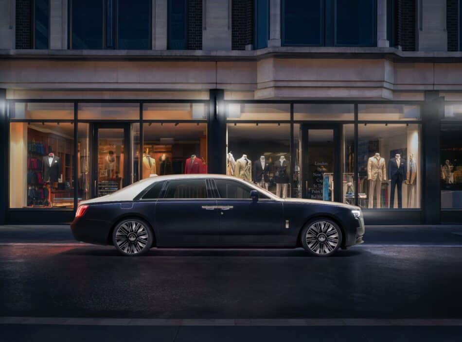 RollsRoyce Ghost Savile Row 1 Watches World Luxury blue sedan parked on a city street in front of a storefront window display with mannequins dressed in suits inside the shop