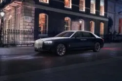 Rolls-Royce Ghost Savile Row Three-quarter front view of a black luxury car (Rolls-Royce) parked on a city street at night, with a brick building and wrought-iron fence in the background.